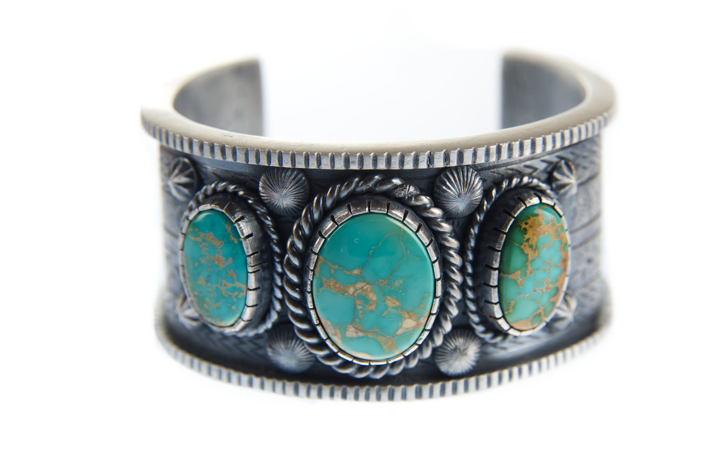 The sterling silver stamped bracelet is lying on its side. This angle features the top of the bracelet which includes three King's Manassa turquoise gemstones encased in bezels. The outside of the bezels are adorned with twisted silver wire. Six sterling silver shot decorations are soldered between the bezels and turquoise cabochons.