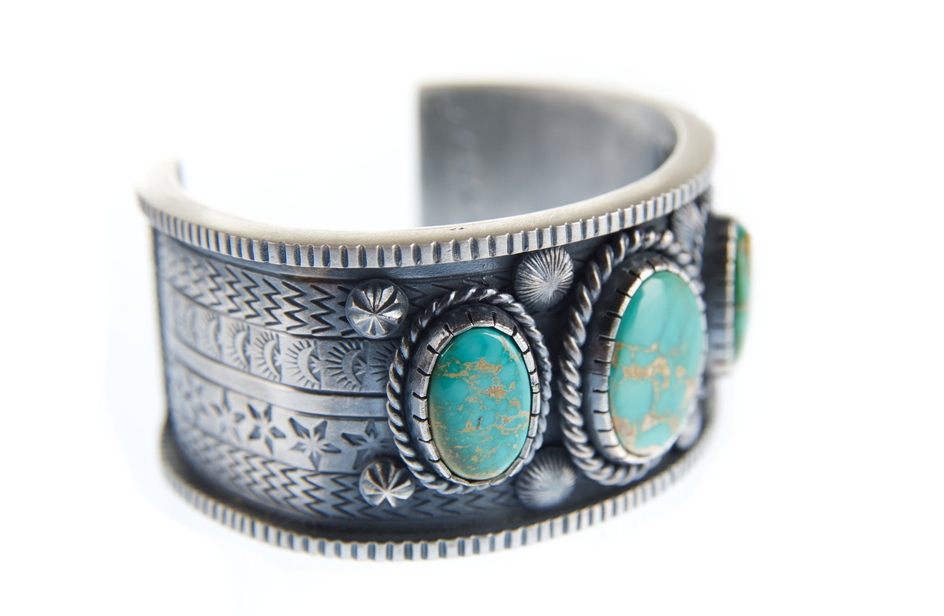 The sterling silver stamped bracelet is lying on its side and positioned so the viewer can see it from a forty-five degree angle. This view highlights the bracelet's texture and the colorful King's Manassa turquoise gemstones.